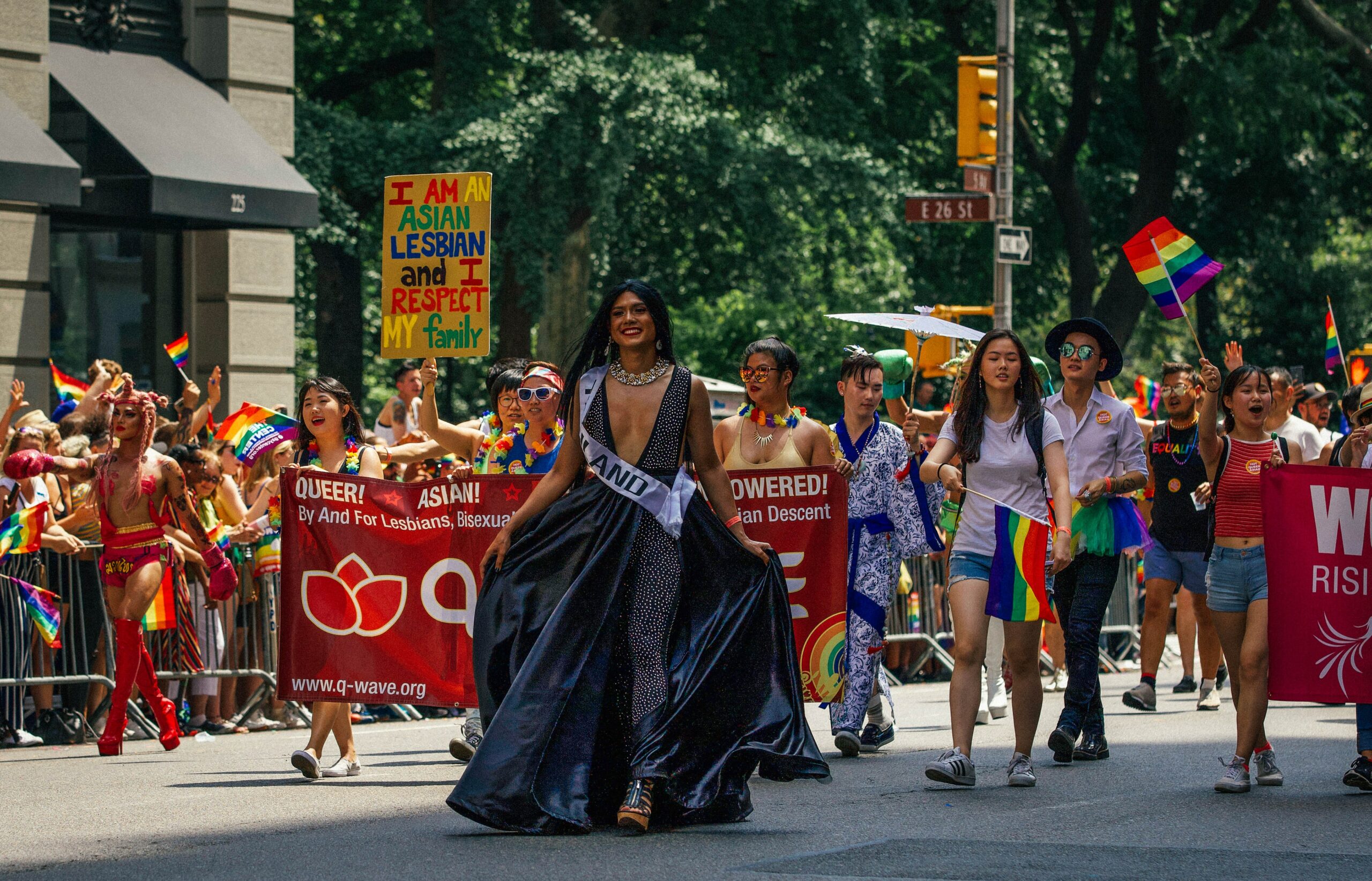 Bild von einem Ausschnitt aus der CSD Parade mit Bunten Flaggen und einem Mann in einem schwarzen Kleid. 