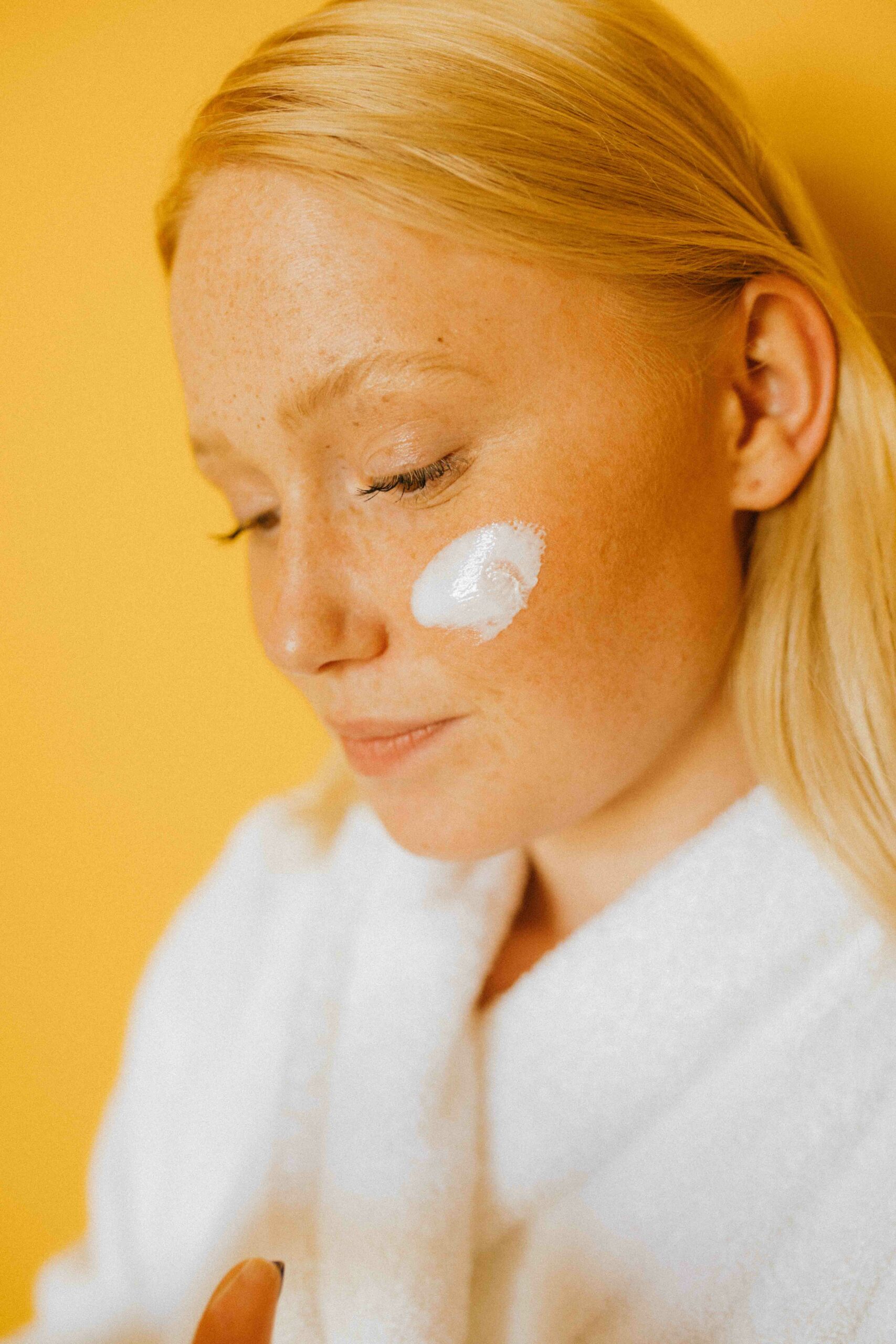 Young woman with long red-blond hair wearing a white bathrobe and has smeared a white cream dot on her left cheek.