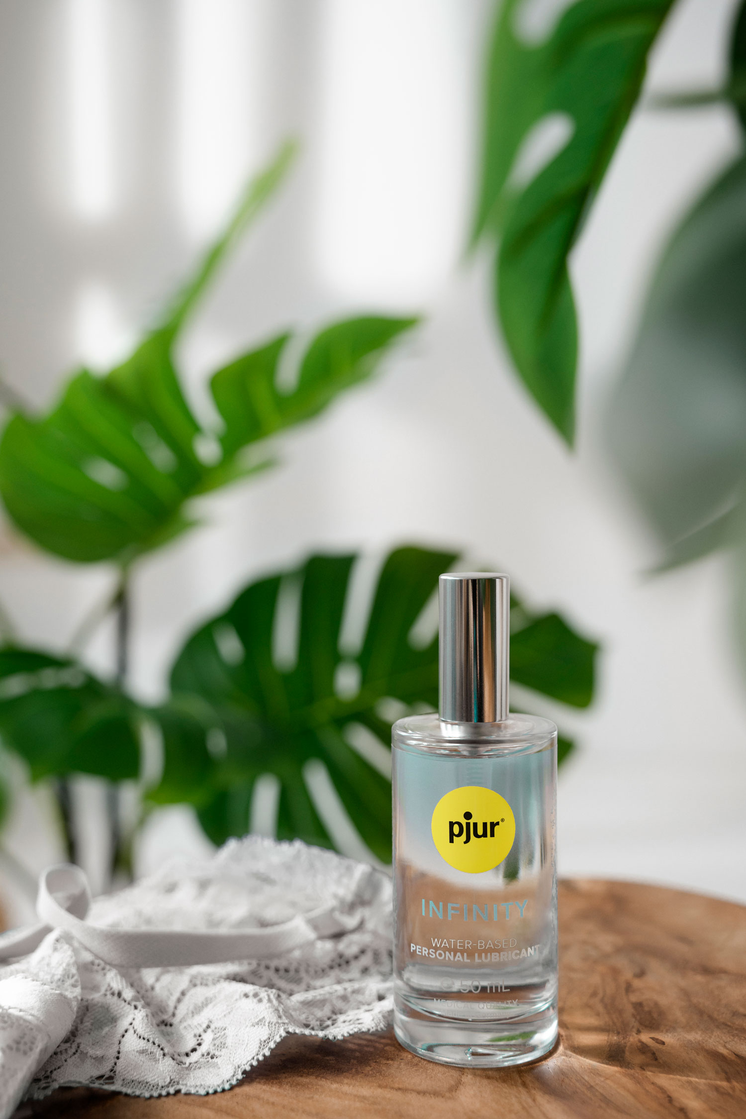 A lubricant in an elegant glass bottle is placed on a wooden table. In the background, there is a green plant, and the room is brightly illuminated. Next to the lubricant, there are also white lingerie.