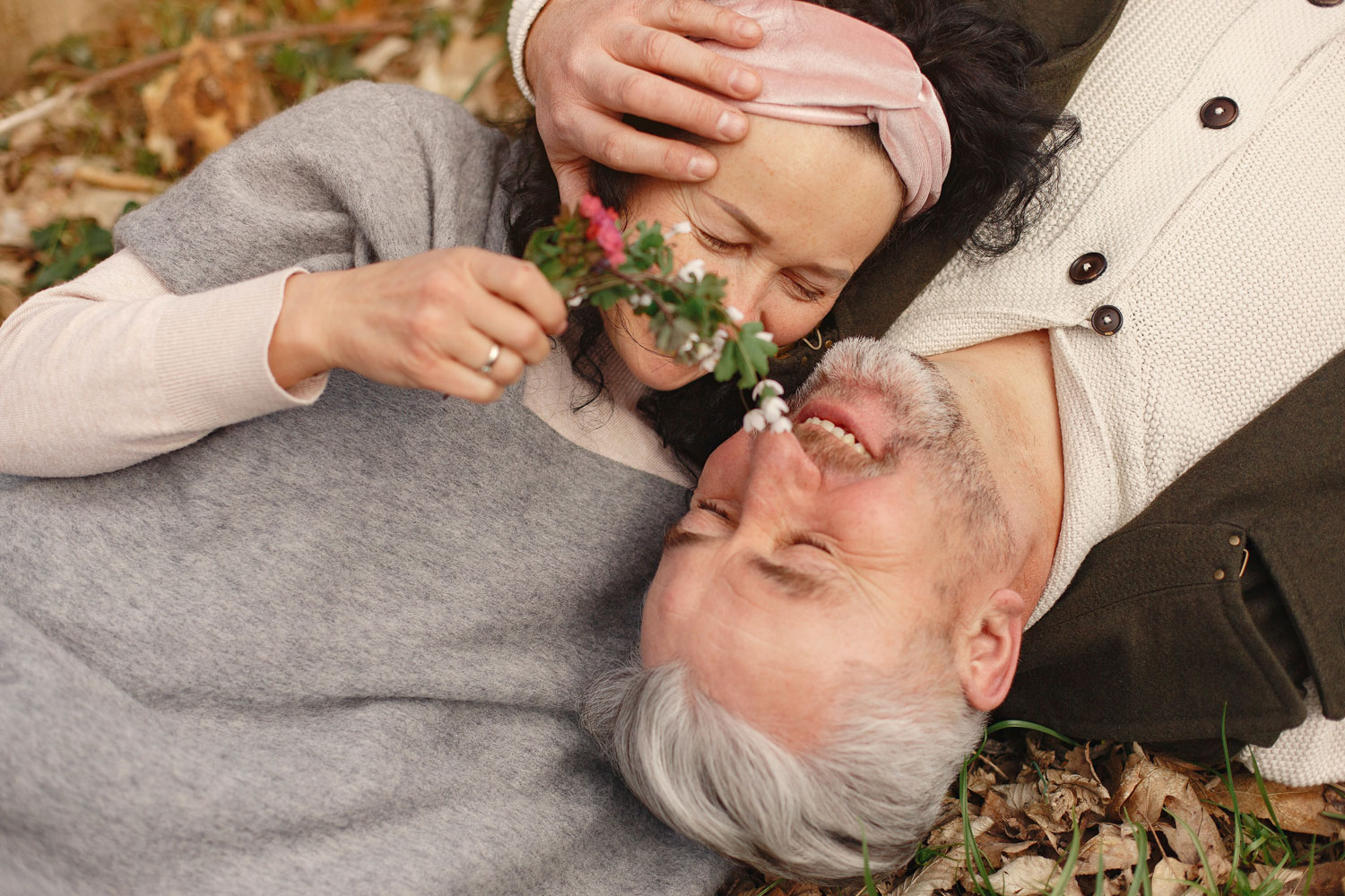 A middle-aged couple lies on the forest floor, cuddling with each other, both smiling at one another.