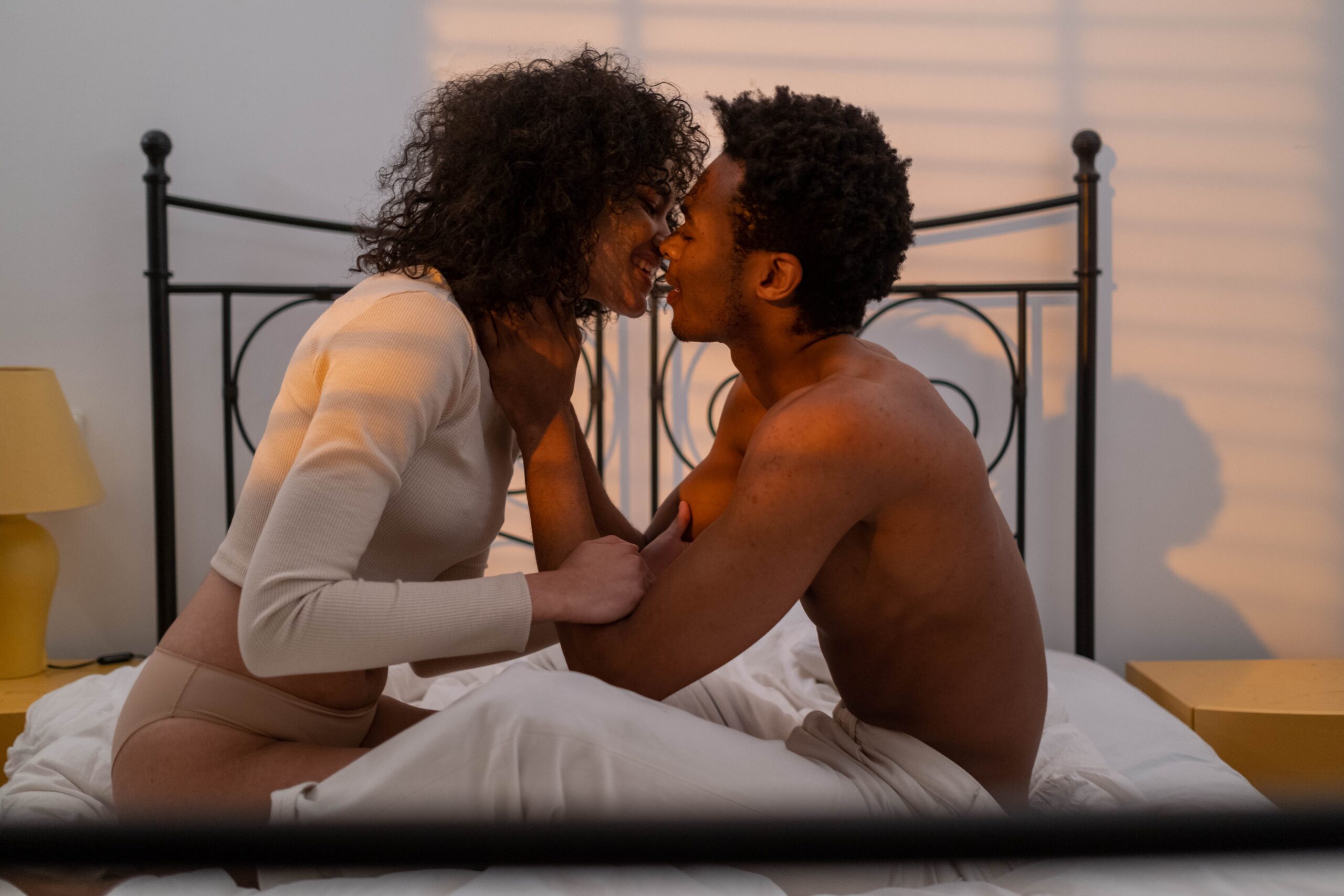 A picture showing a smiling couple sitting on a bed and kissing.