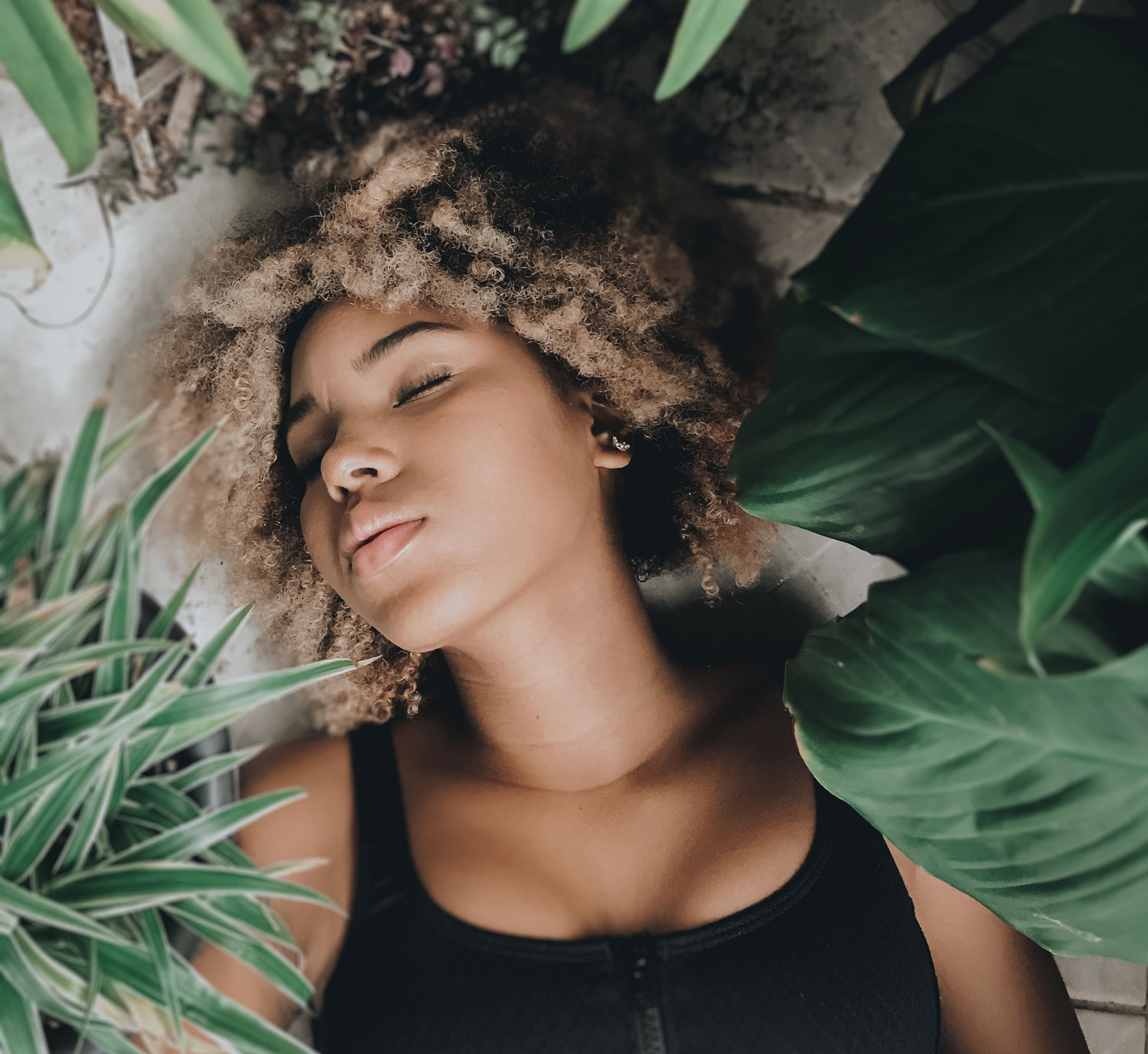 The image shows the upper body of a woman lying relaxed among some plants. Her eyes are closed and she is relaxing on the ground.