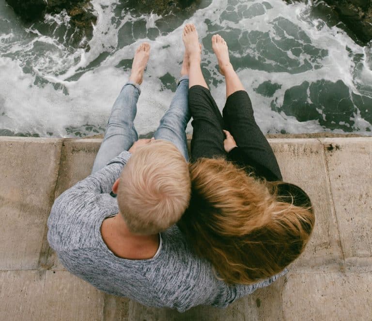 A young man and a young woman sit together on the seashore. Their heads are pressed together and the man has his arm around the woman's waist. The photograph is taken from above.