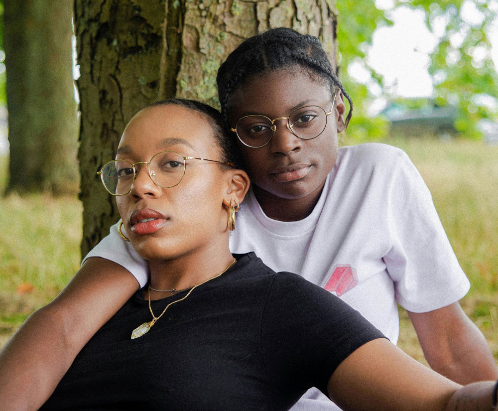 A young female couple sit together. One of the women has her arm draped over the other woman's shoulder.