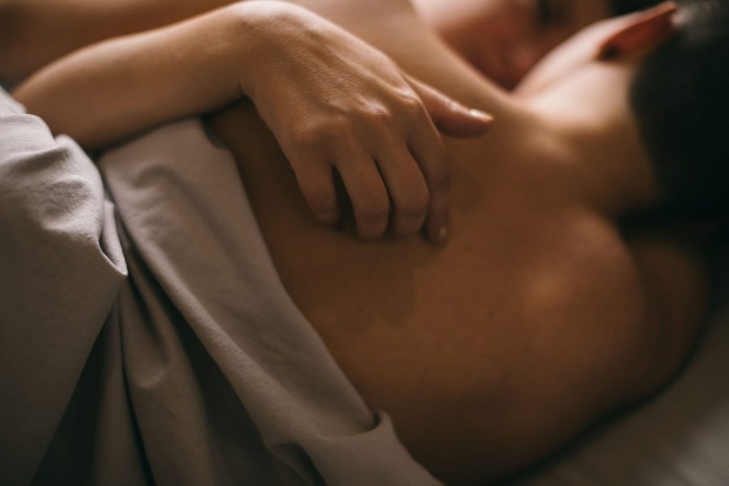 A couple lie embracing in bed, naked under the sheets. One of the partners gently rests their hand on the other partner's back.