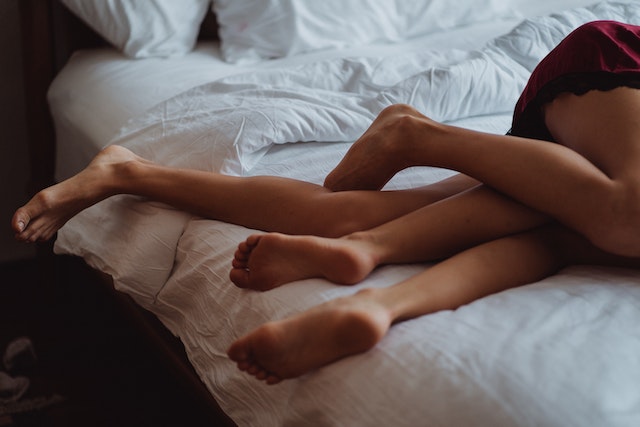 A couple lie on a bed with white sheets. Only their legs are visible; the heads are not pictured. Their legs are entwined.