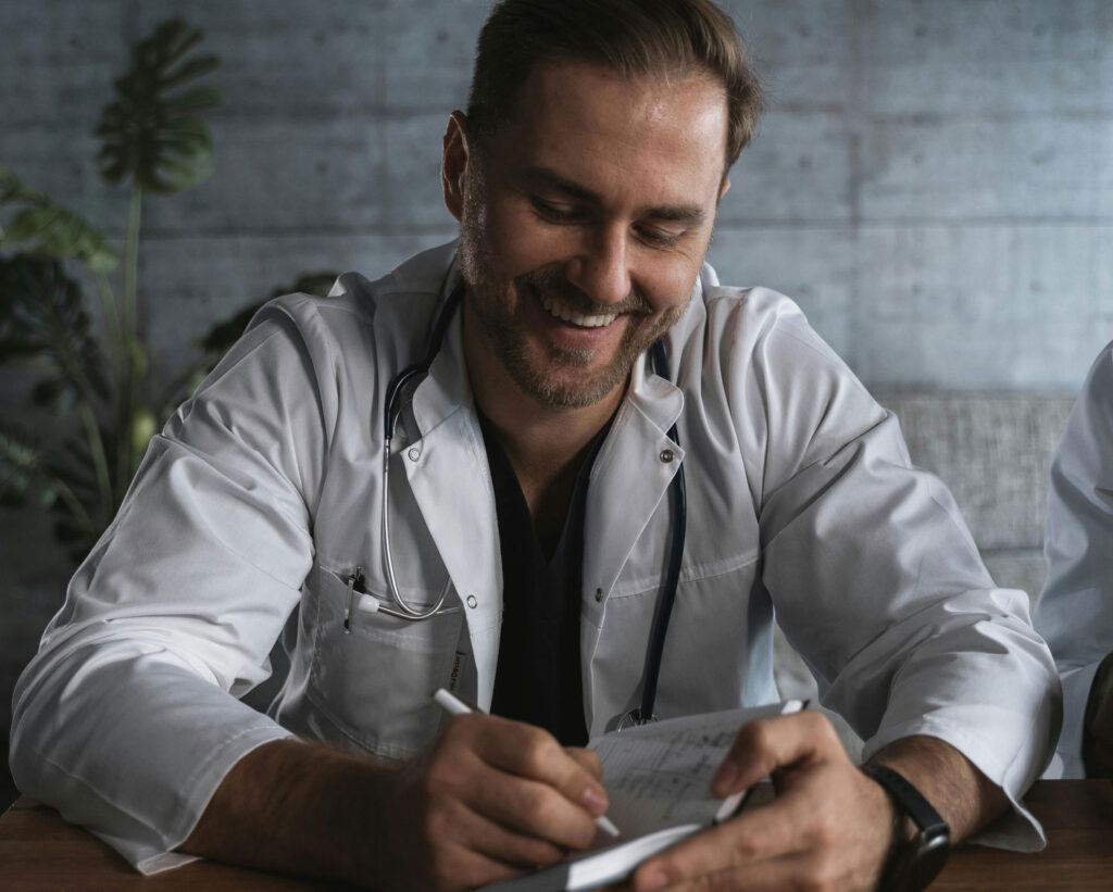 Smiling doctor making notes at a table with a white coat and stethoscope.